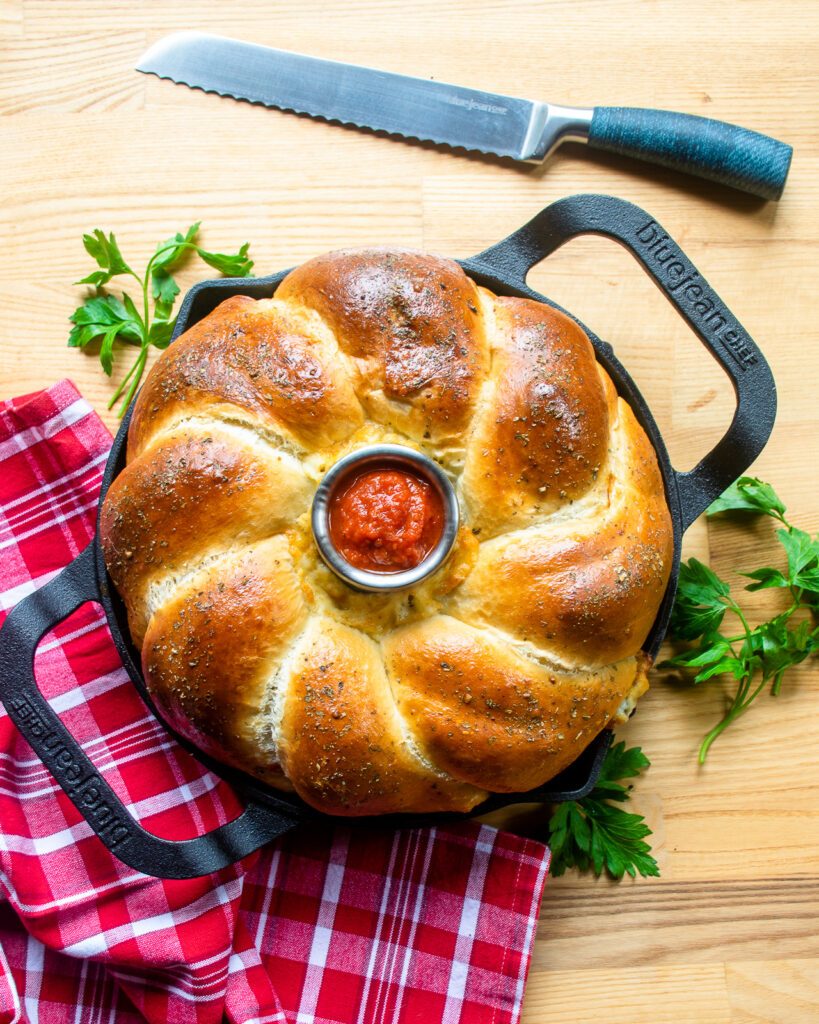 Looking straight down on a bread ring in a cast iron pan with pizza sauce in the middle, next to a bread knife and a red and white kitchen towel.