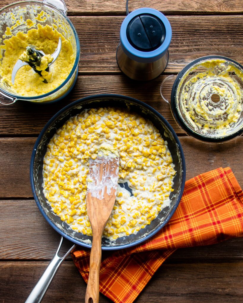 Cream and corn in a skillet with a wooden spoon on a wooden table, next to a chopper with puréed corn inside and an orange towel nearby.