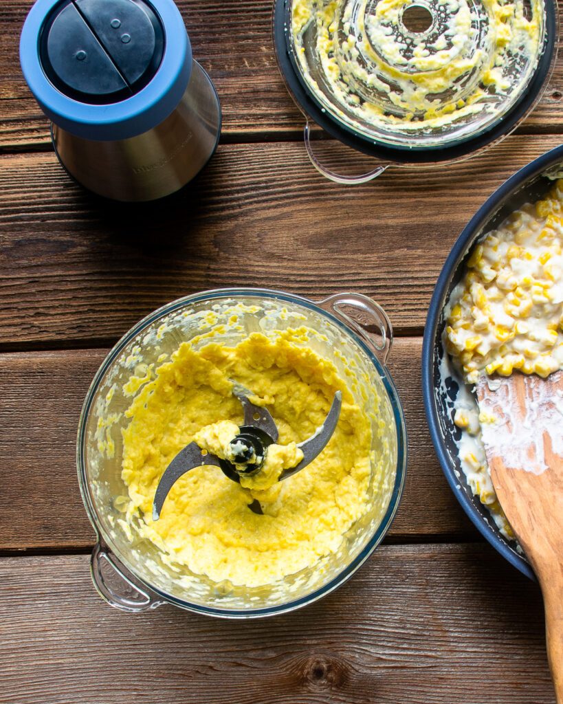 Looking down into a chopper with puréed corn inside and a skillet of creamy corn next to it.