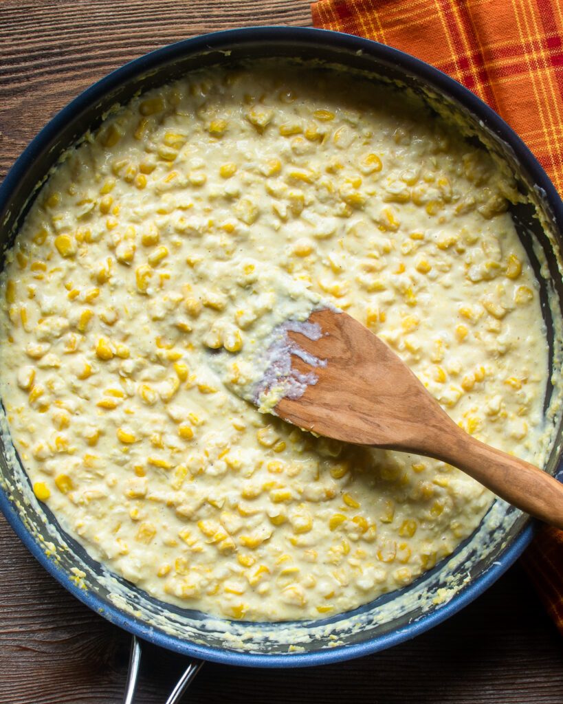 Looking down into a skillet of creamed corn with a wooden spoon and an orange hand towel next to it.