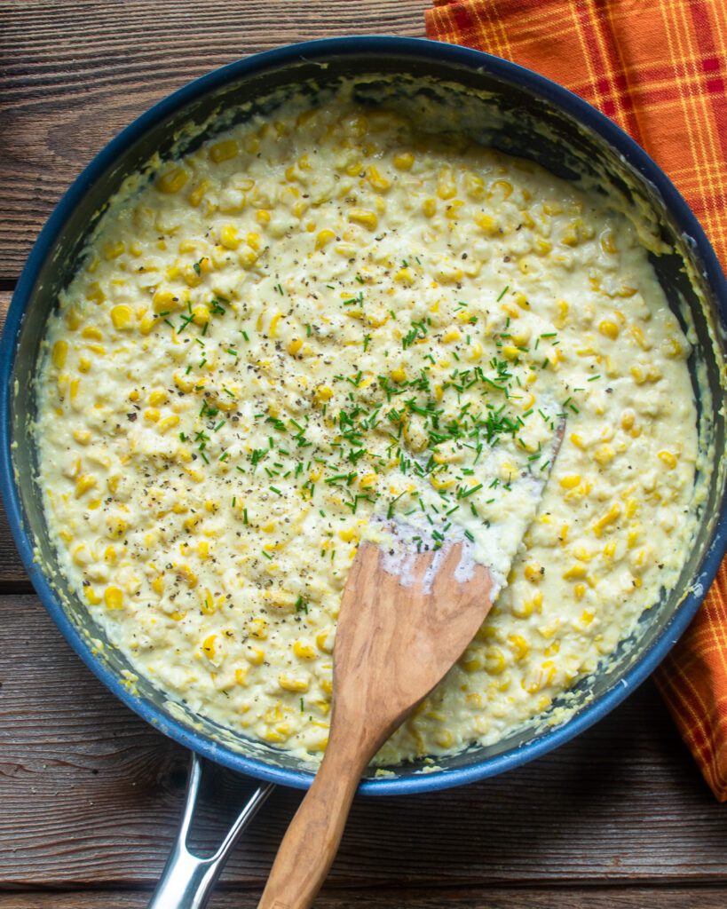 A skillet of creamed corn with chives and black pepper being stirred in with a wooden spoon.