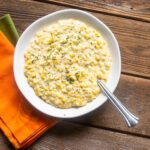 Creamy corn in a white bowl with a silver spoon next to orange and green napkins.