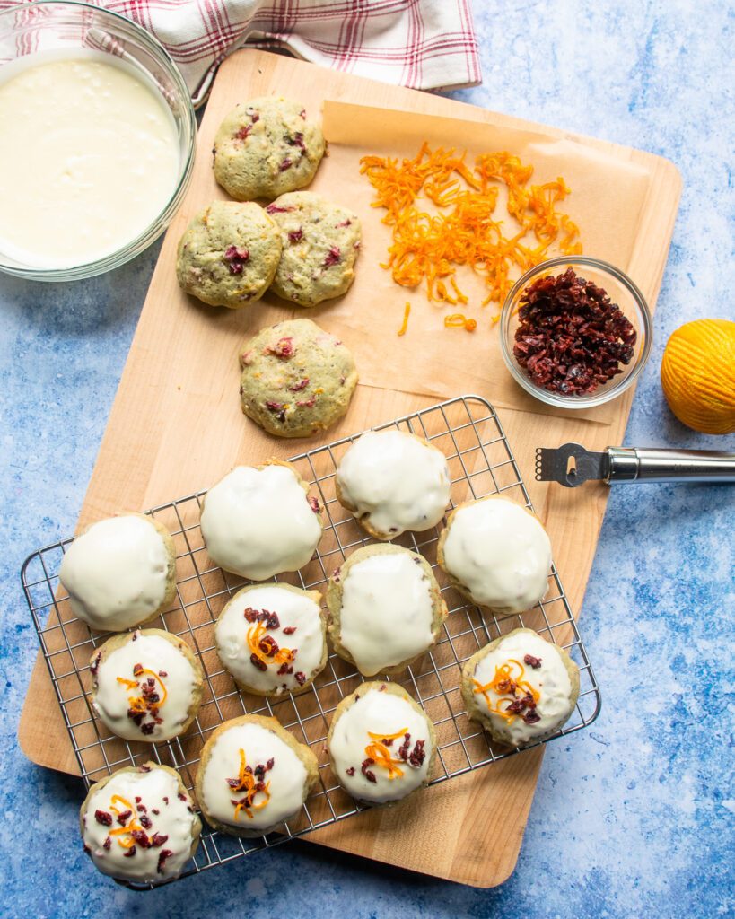 Looking down on a cutting board with some unfrosted cookies, a bowl of frosting and some frosted cookies on a cooling rack.