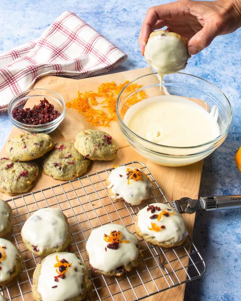 A hand dipping a cookie into a bowl of frosting with partially frosted cookies on a cooling rack in front.