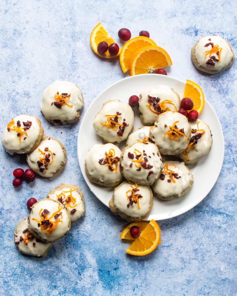 Orange Cranberry Frosted Cookies on a plate on a blue countertop with orange slices and fresh cranberries around.