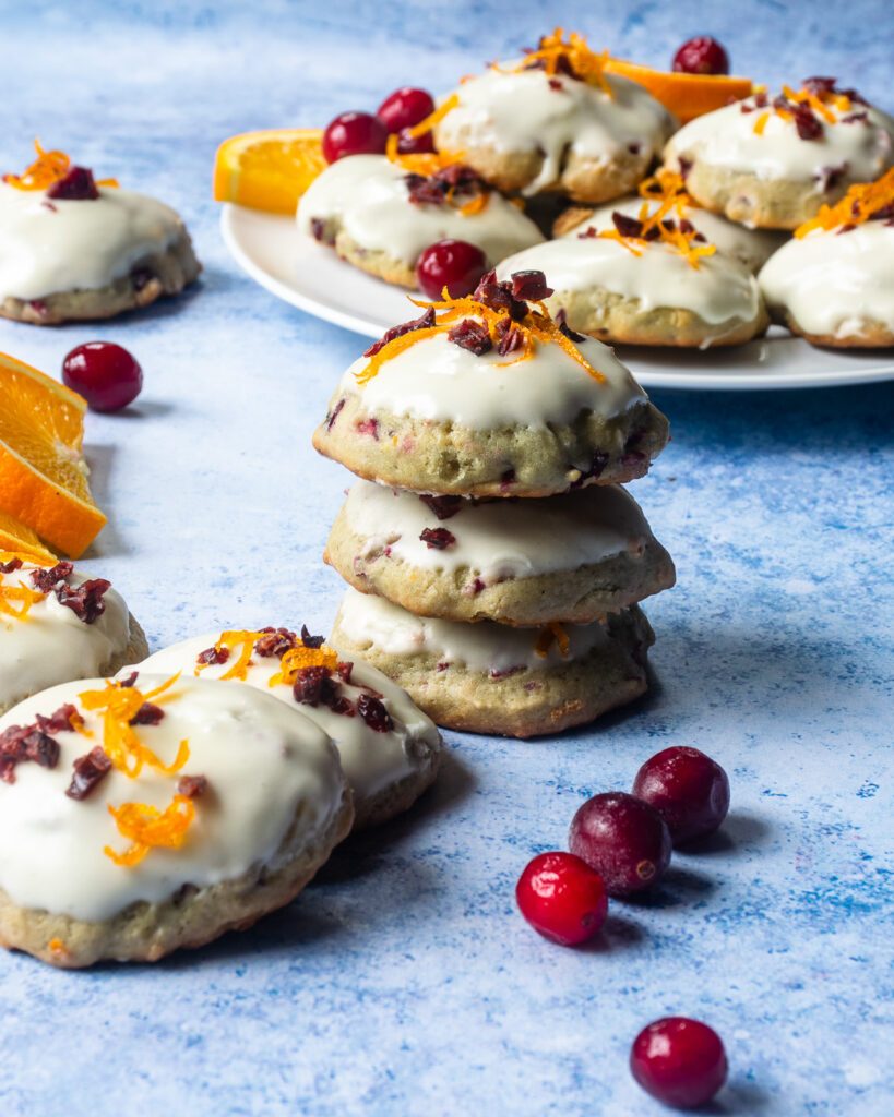 A stack of three orange cranberry frosted cookies with a plate behind it as well as more cookies, orange slices and fresh cranberries around.