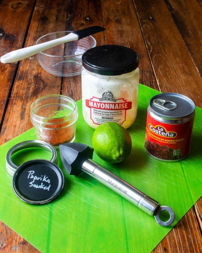 Ingredients on a green mat on a wooden table - chipotle peppers, mayonnaise, smoked paprika, lime, reamer, bowl and spatula.
