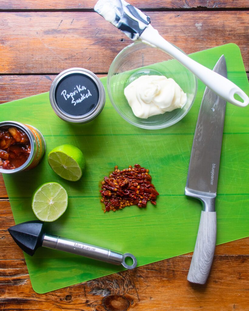 Looking down on chopped chipotle in adobe next to a halved lime, a bowl of mayo and a jar of smoked paprika.