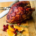 A cranberry glazed roast turkey breast on a cutting board with rosemary, orange and cranberries.