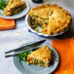 A grey plate with a pie of vegetable pie, next to orange napkins and the rest of the whole pie in the background.