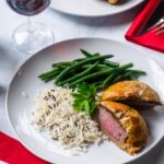 Individual Beef Wellington on a white plate with rice and green beans and a table setting with a red napkin and red wine.