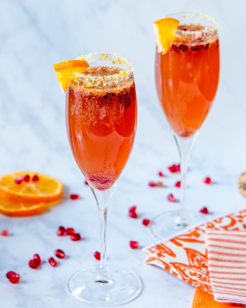 Two champagne flutes of pomegranate tangerine champagne cocktail on a marble counter with pomegranate seeds adn a few slices of tangerine scattered around.