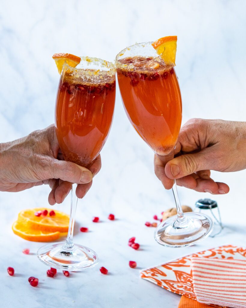 Two hands clinking champagne flutes filled with a pomegranate tangerine champagne cocktail.