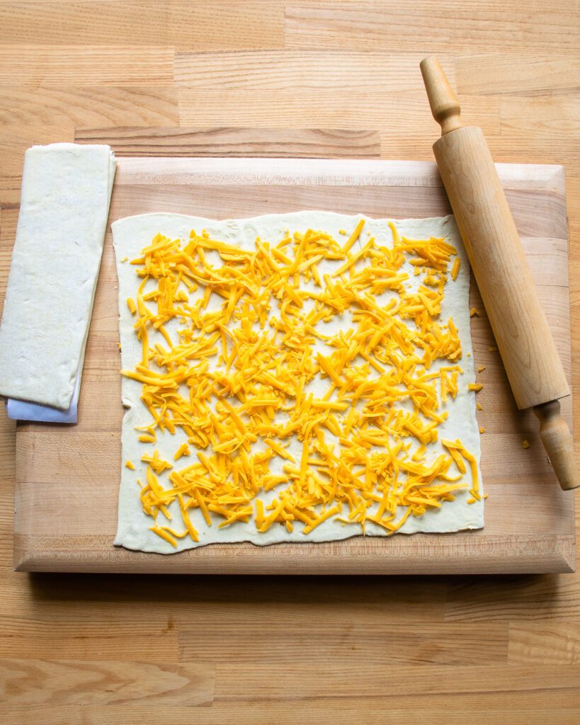 A sheet of puff pastry with Cheddar cheese all over it on a wooden cutting board, and a rolling pin next to it.