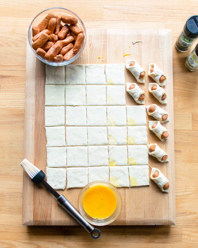 A sheet of puff pastry cut into 36 squares with cocktail wieners and egg wash - the process of making pigs in a blanket.