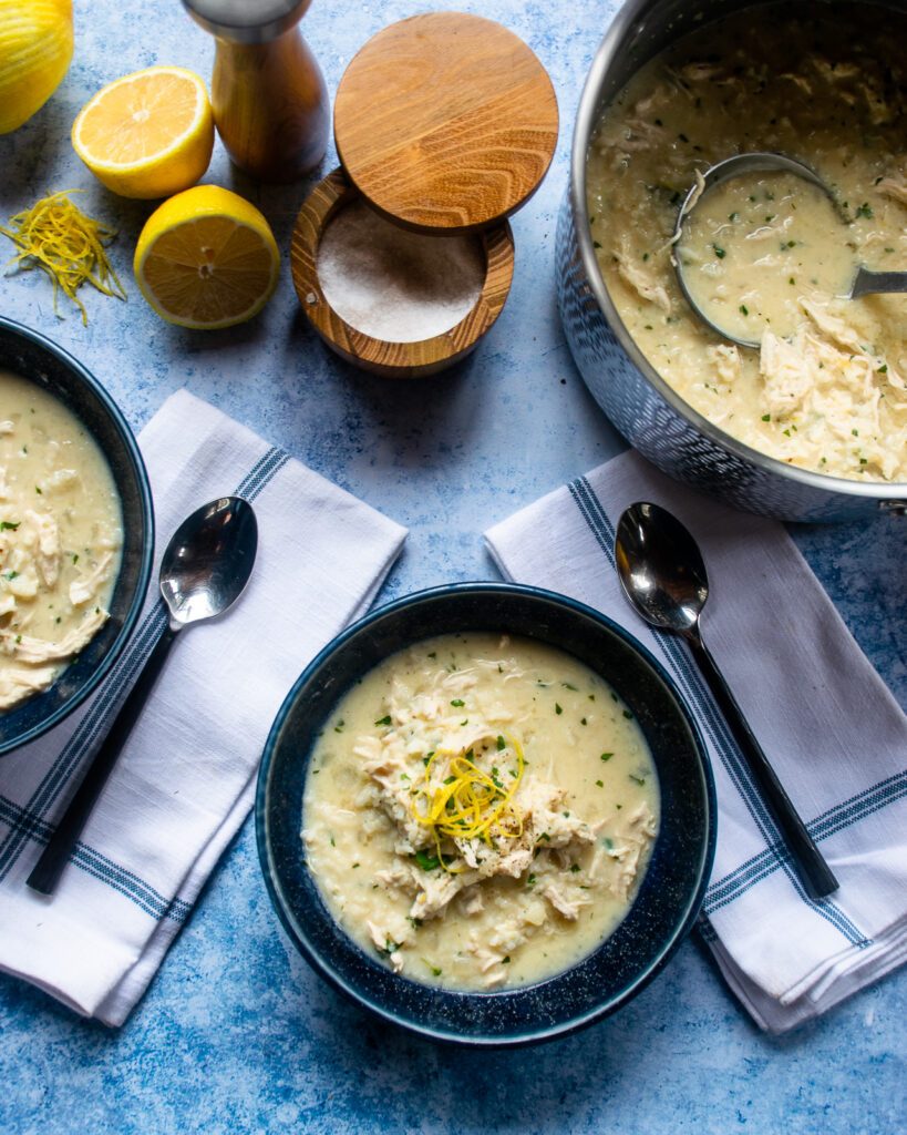 Two bowls of avglolemono soup with a stockpot of soup, salt, pepper and extra lemon near by.