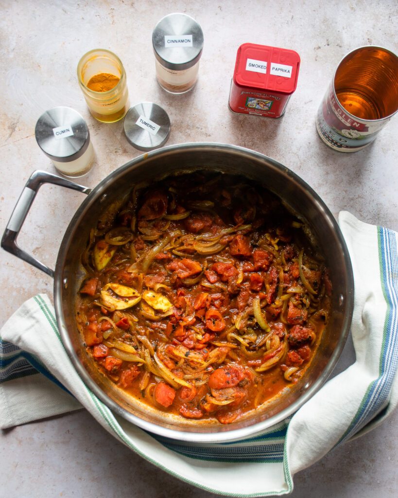 A spiced tomato sauce in a saute pan with spice jars next to it.