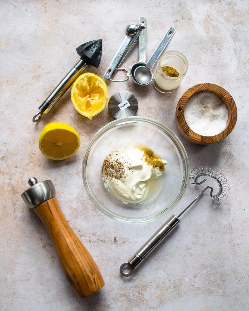 A bowl of sourcream with other ingredients and a whisk, peppermill, salt cellar, measuring spoons and a reamer near by.