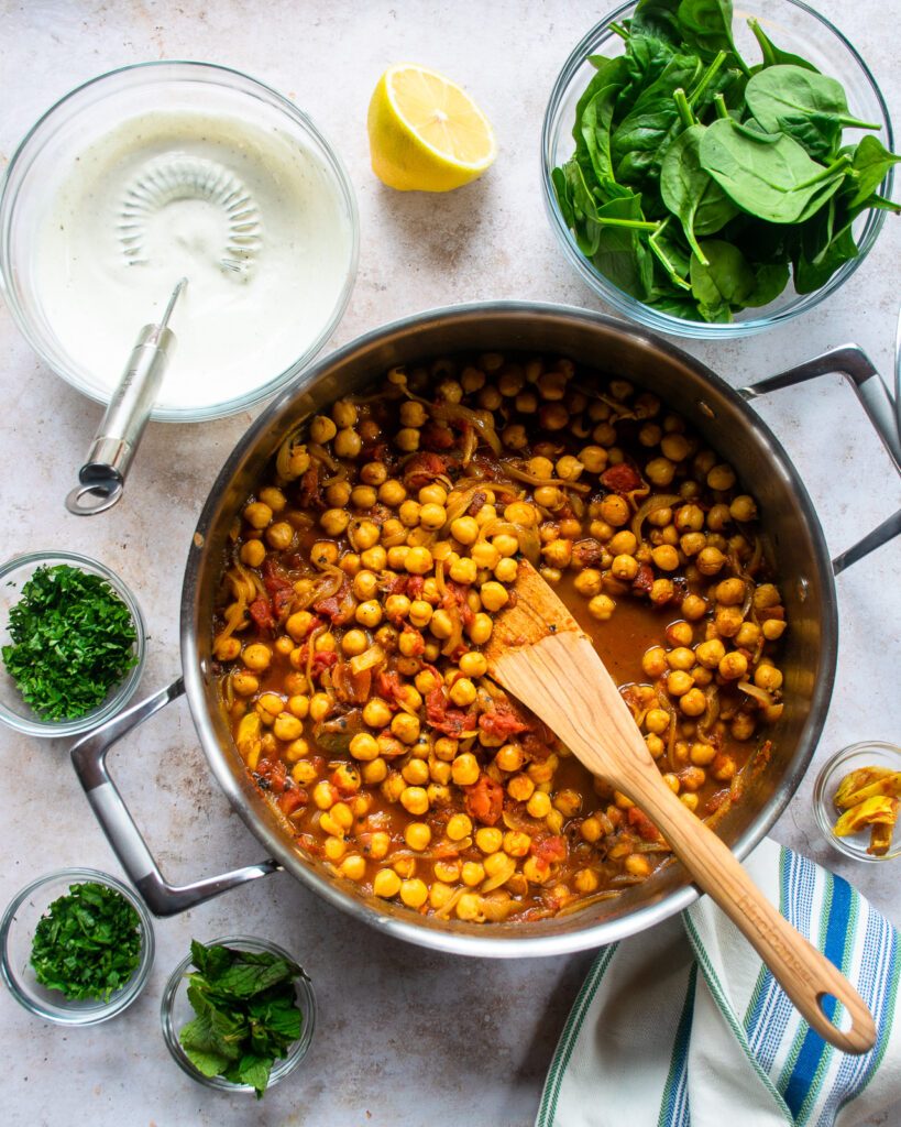 A saute pan with braised chickpeas and a wooden spoon sticking out of it, with herbs, spinach a lemon and a bowl of sour cream next to it.