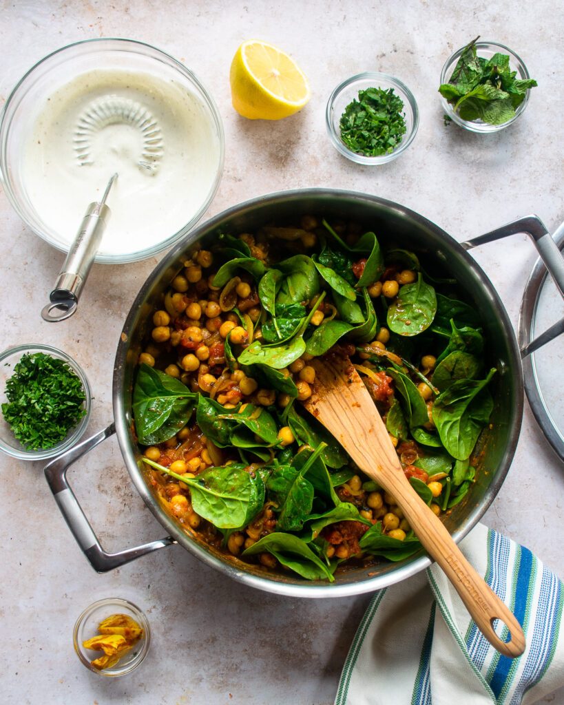 A saute pan of braised chickpeas with spinach being stirred into the pan and some other ingredients on the counter near by.