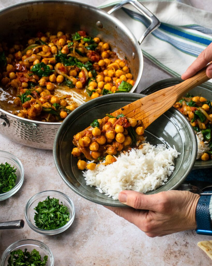 Hands spooning braised chickpeas into a green bowl with rice in it.