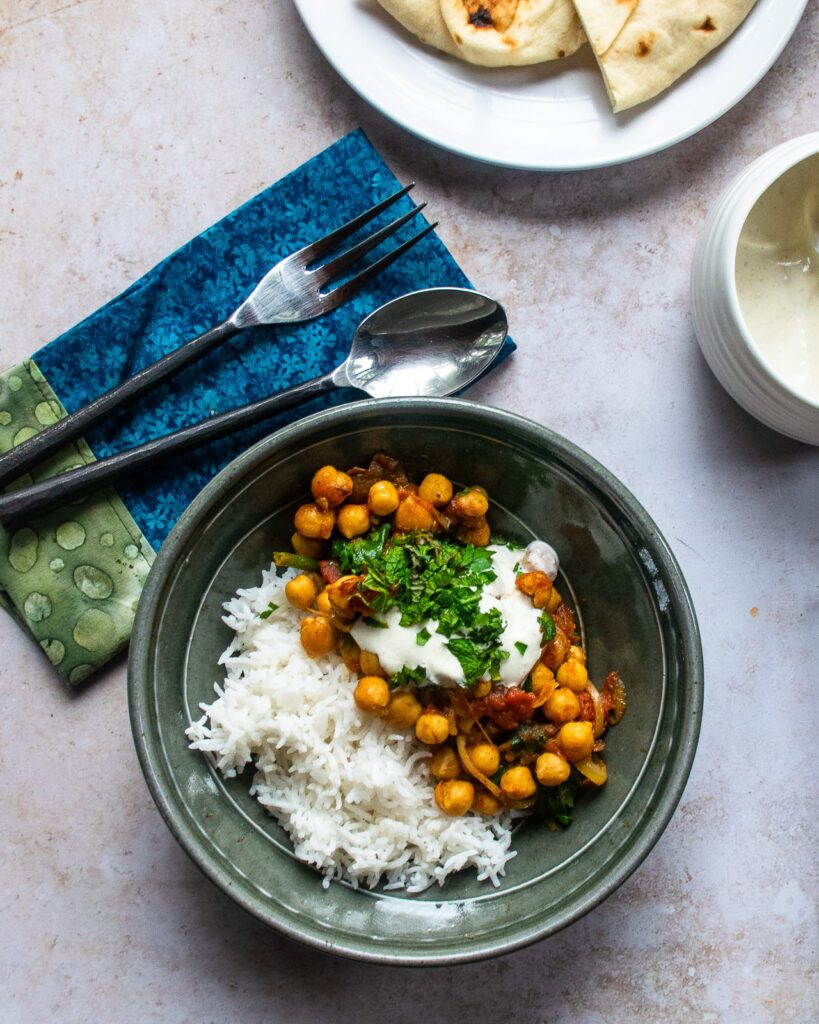 Looking straight down into a green pottery bowl of braised chickpeas with rice, a place setting a bowl of white sauce and a plate of naan.