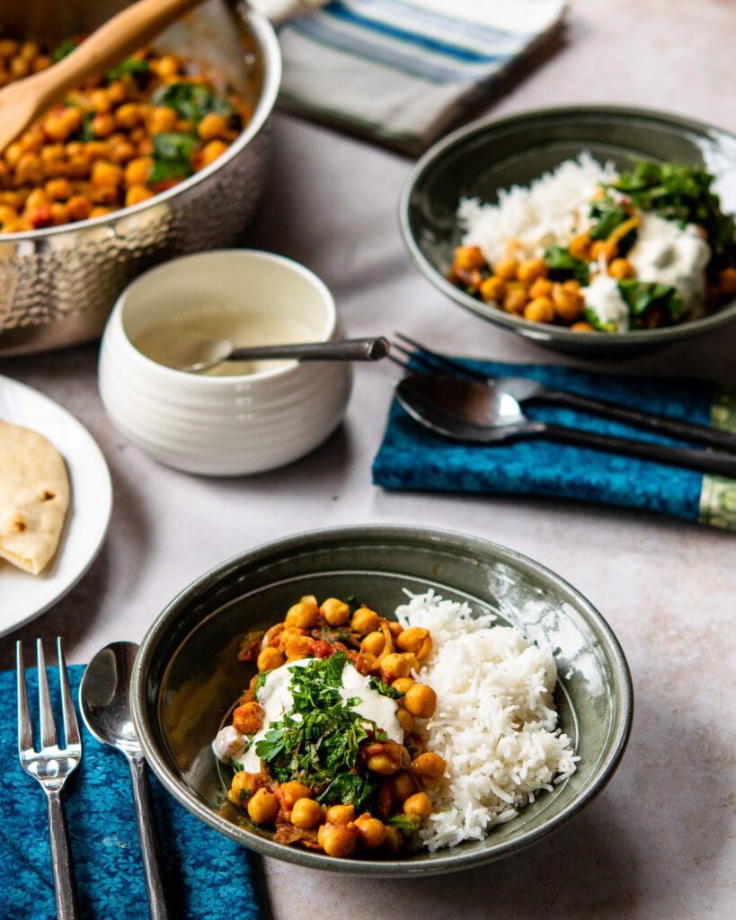 Two green pottery bowls with tomato braised chickpeas and rice, place settings a bowl of sauce and a saute pan with more chickpeas in it.