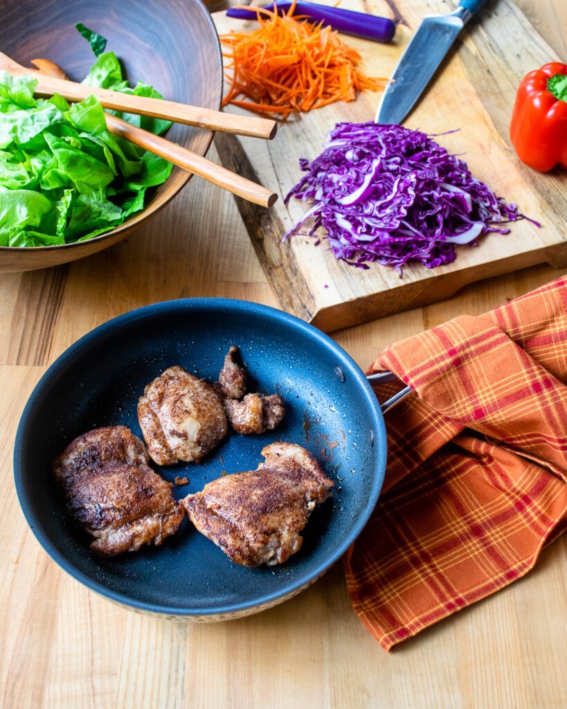 Chicken thighs cooked in a skillet with a cutting board of chopped vegetables and a bowl of lettuce in the background.