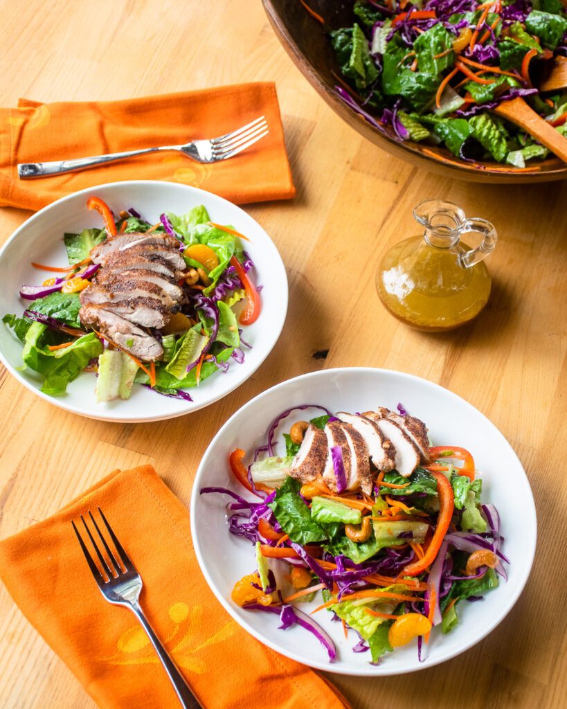 Looking at two white bowls with Chinese Five Spice Chicken Salad, orange napkins and a larger bowl of salad in the background.