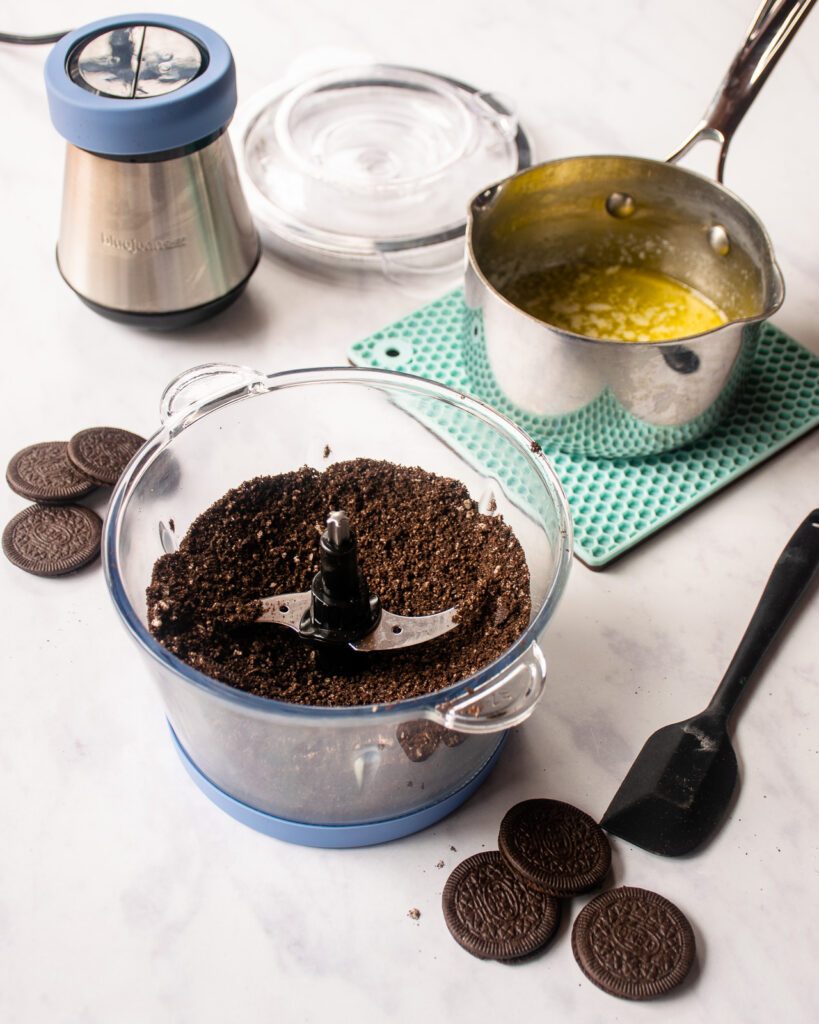 A chopper with cookie crumbs inside, with a saucepan of melted butter behind it.