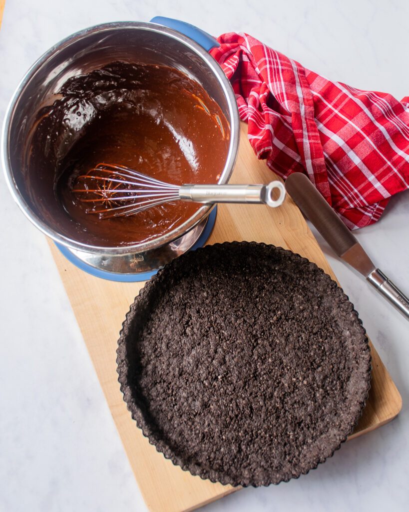 Looking down on a chocolate tart crust and a bowl of chocolate ganache with a red and white kitchen towel near by.