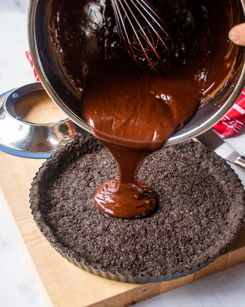 Chocolate being poured from a stainless steel bowl into a chocolate tart crust.