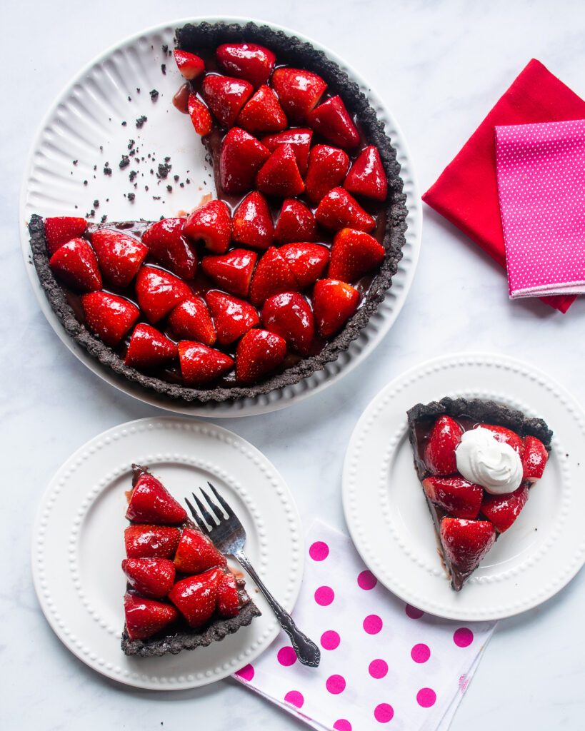 A large chocolate strawberry tart on a large white plate with two smaller plates, each with a slice of the tart.
