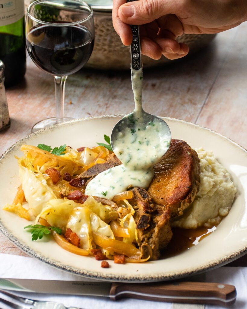 A ladle pouring some parsley sauce on some braised pork chops with cabbage and bacon.