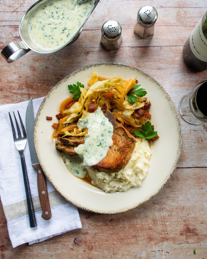 Pork chops with cabbage and bacon on a plate on a wooden table with a gravy boat of parsley sauce and place setting.