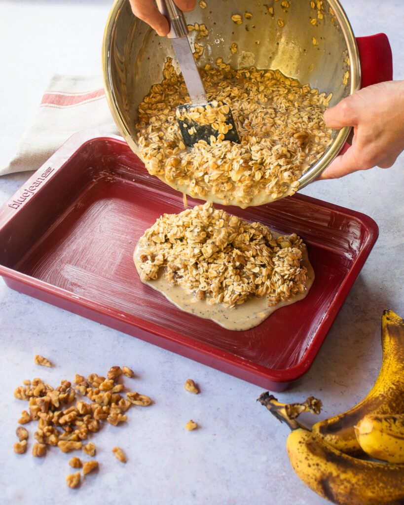 Pouring an oatmeal mixture from a bowl into a red baking pan.