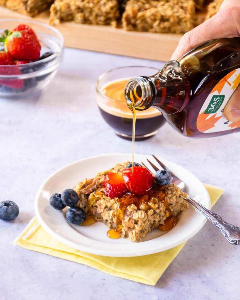 Syrup being poured on a baked banana oatmeal square with berries and a coffee nearby.
