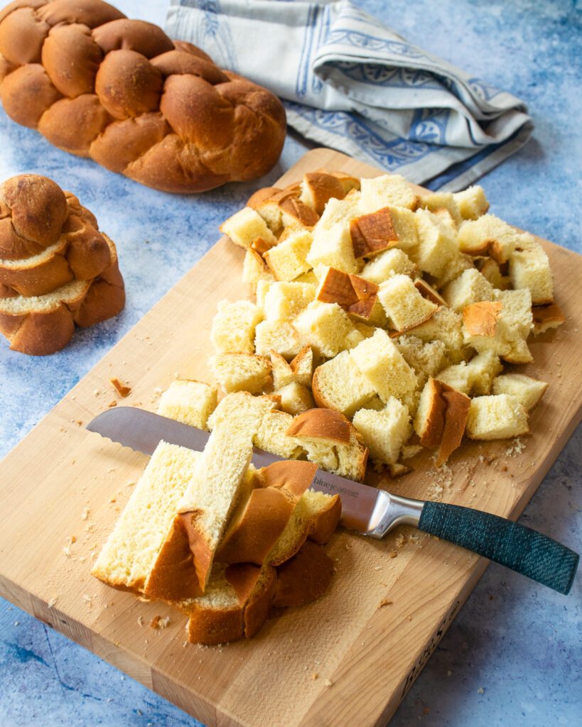 Challah bread being cut into cubes with a blue jean chef bread knife on a cutting board with a blue countertop.