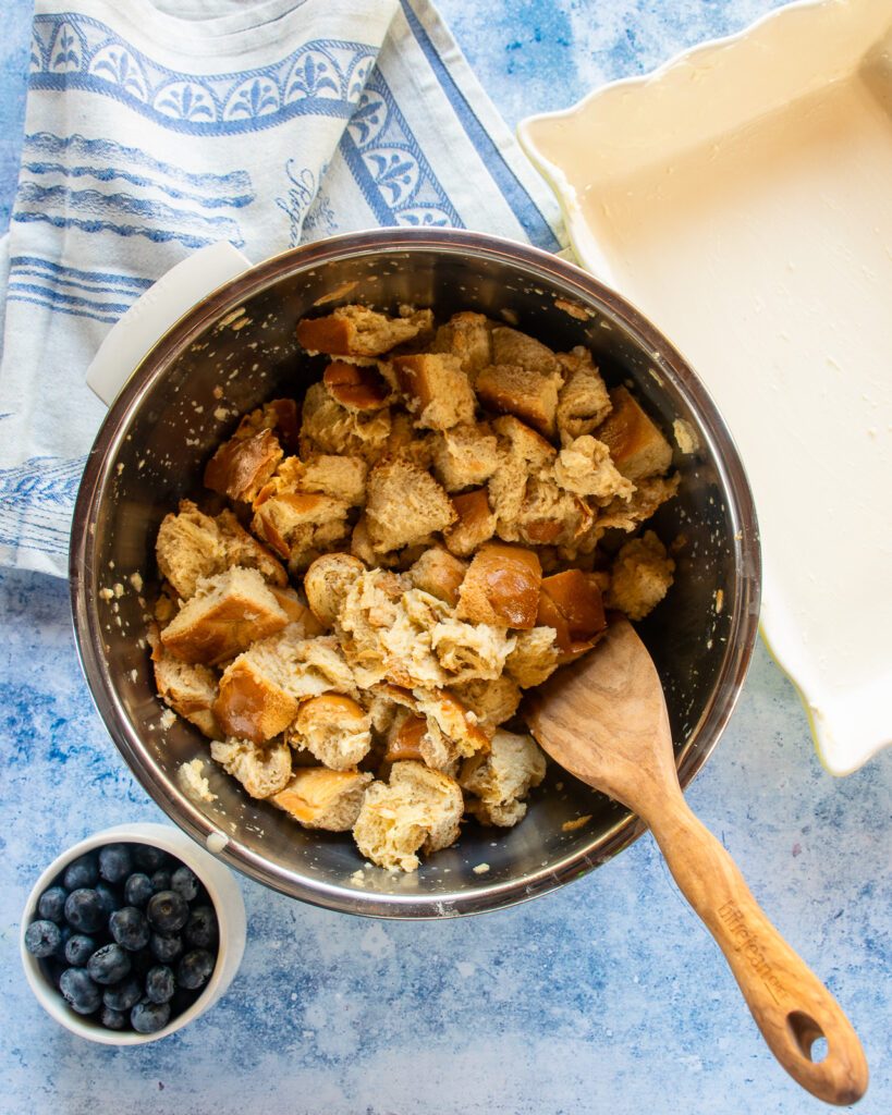 Bread cubes in a bowl of egg custard with a wooden spoon sticking out of it, a small bowl of blueberries and a ceramic baking dish near by.