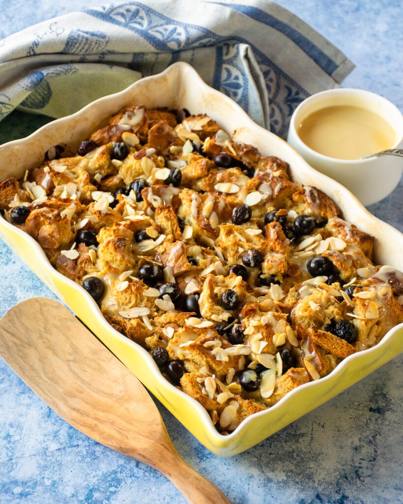 blueberry bread pudding in a yellow ceramic baking dish with mascarpone sauce and a wooden spoon next to it.