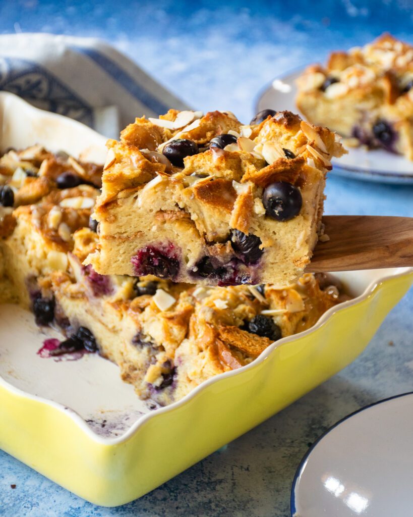 A wooden spoon lifting a piece of blueberry bread pudding out of a baking dish.