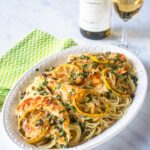 A platter of chicken piccata on thin spaghetti with a glass and bottle o white wine in the background.