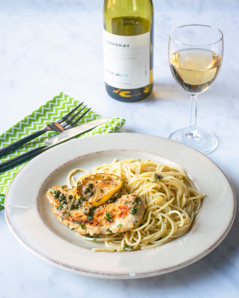 A white plate of chicken piccata on thin spaghetti with a glass and bottle of white wine and green napkins.