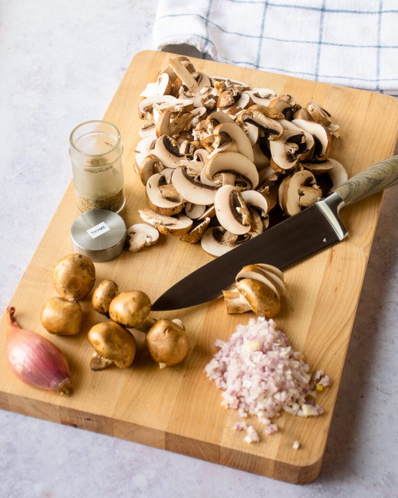 Sliced mushrooms and minced shallots on a cutting board with a knife.