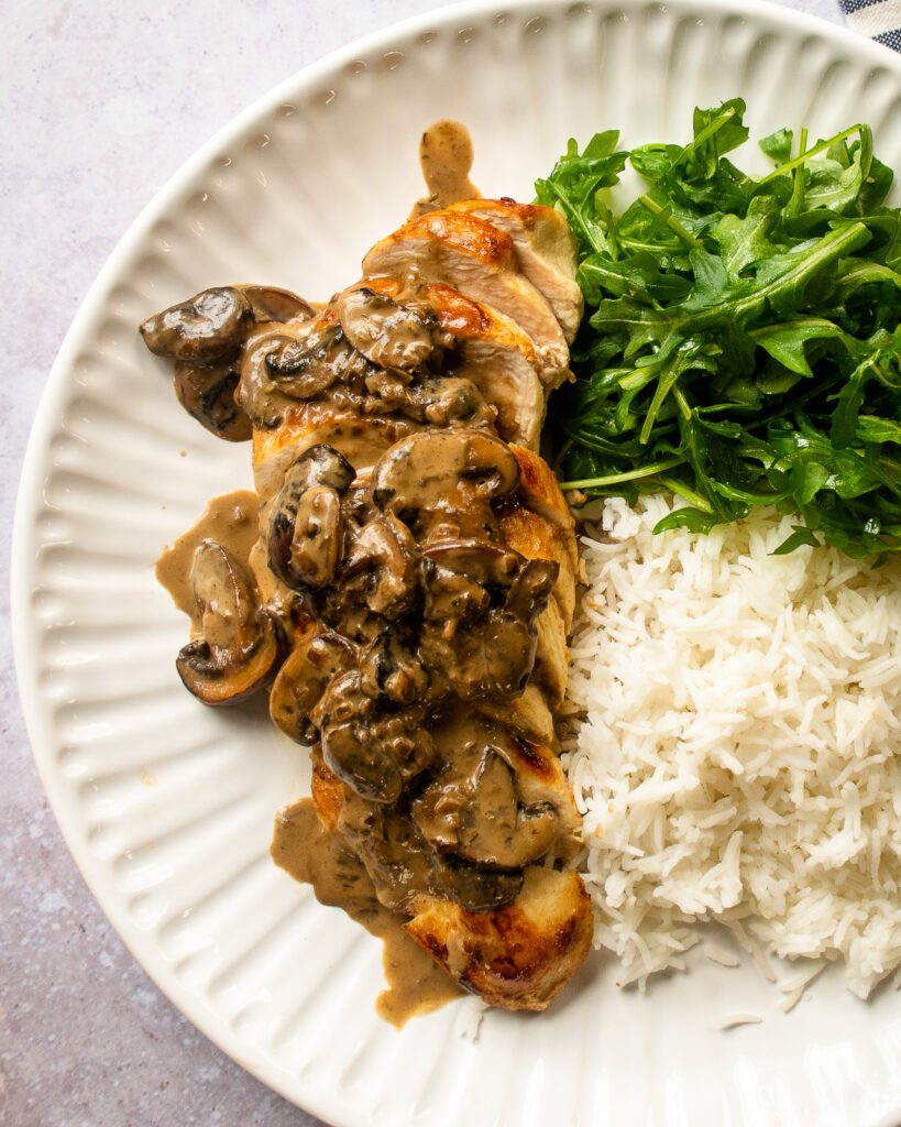 Chunky mushroom Sauce on a chicken breast with rice and salad greens, on a white plate.