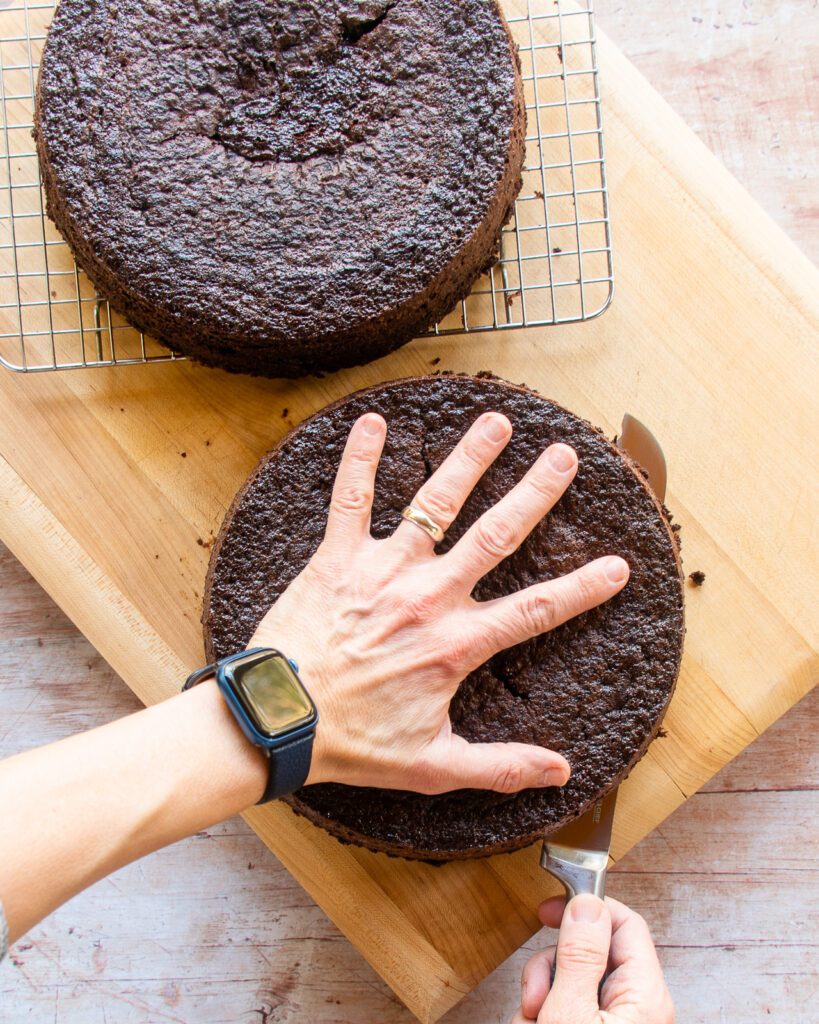 Hands cutting a chocolate cake layer in half horizontally on a wooden cutting board.