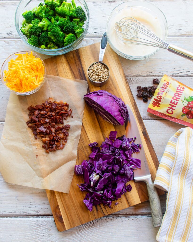 Ingredients on a cutting board: cooked bacon, chopped cabbage, blanched broccoli, dried cherries, sunflower seeds, cheddar cheese and salad dressing.