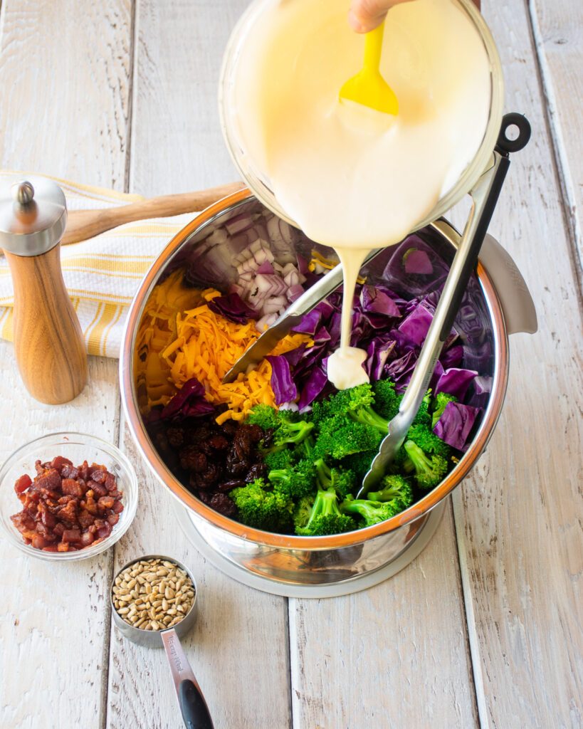 Pouring salad dressing into a bowl with vegetables in it and some dried fruit and seeds on the table.
