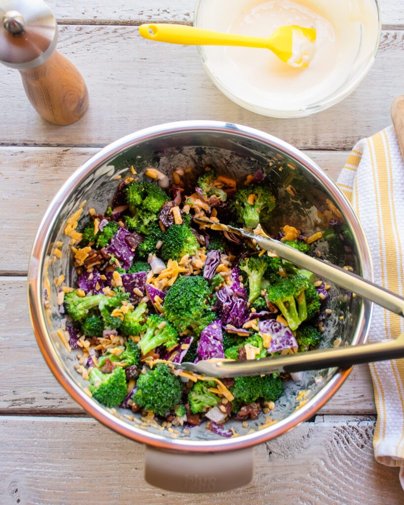 Broccoli salad tossed together in a stainless steel bowl with tongs in it.