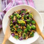 Broccoli Salad in a white bowl on a white wooden table with salad servers and a purple striped towel.